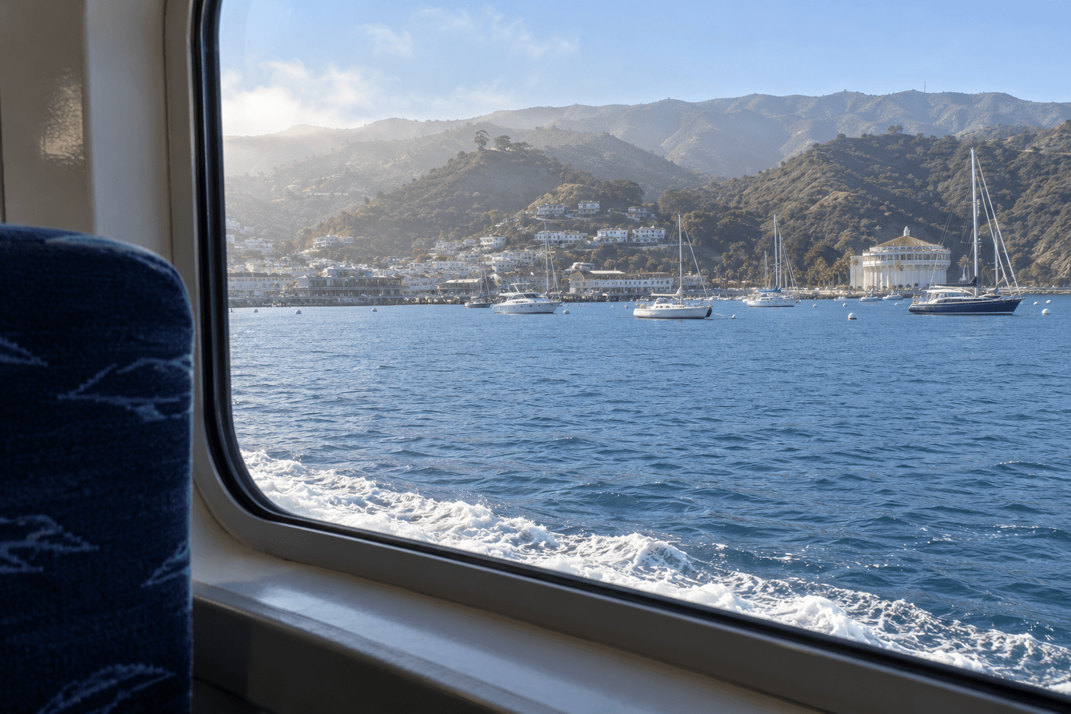 View through a passenger-ferry window toward an island harbor with sailboats and hazy hills